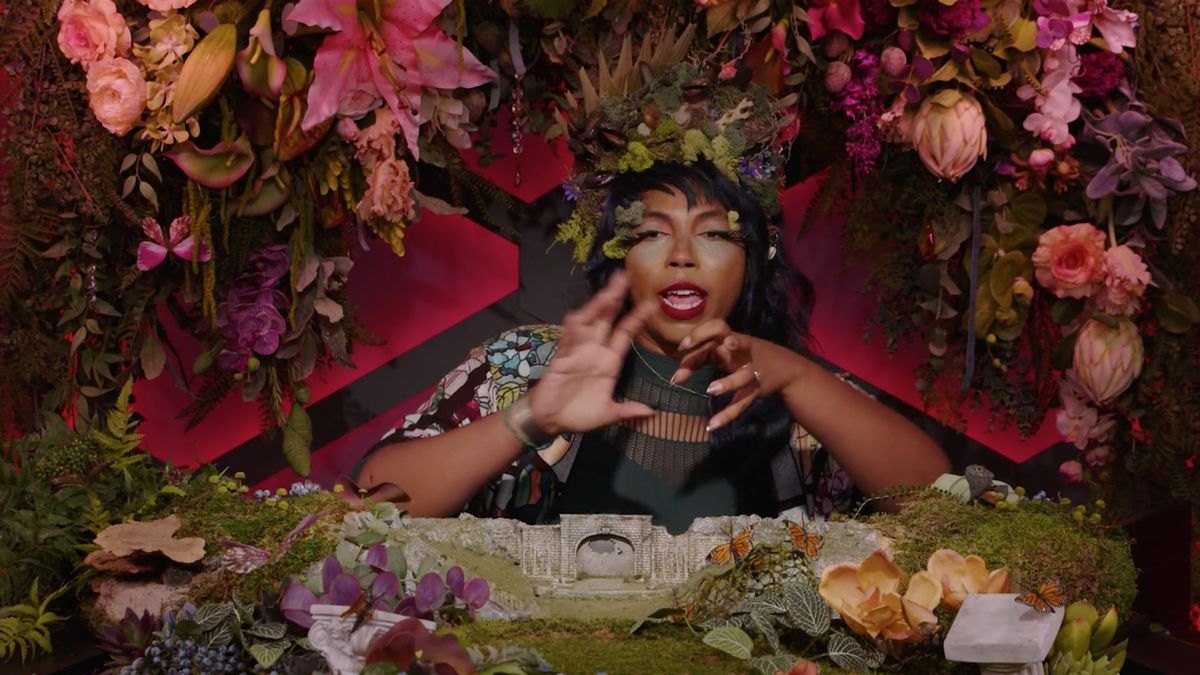 Aabria Iyengar pantomima mentre tesse un incantesimo. Sotto di lei c'è un diorama in miniatura di un campo verde con rovine. Intorno alla sua miriade di fiori di seta occludono la cornice.