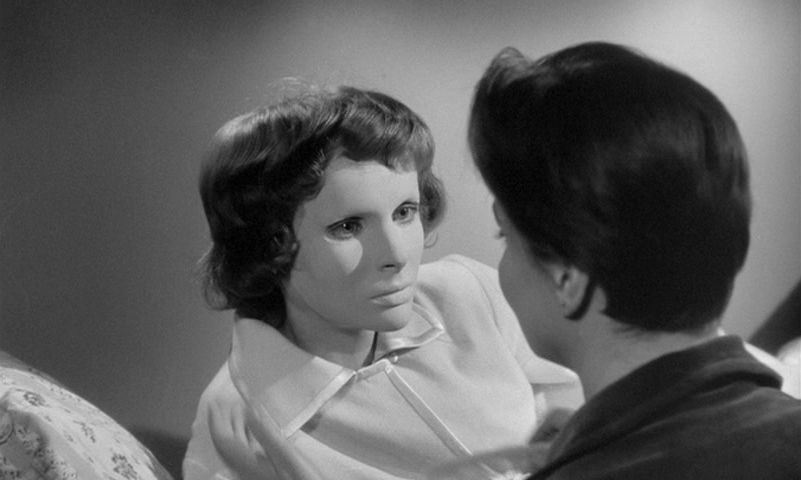 Edith Scob indossa la sua maschera bianca in Eyes Without a Face mentre si appoggia su un letto e parla con qualcuno.