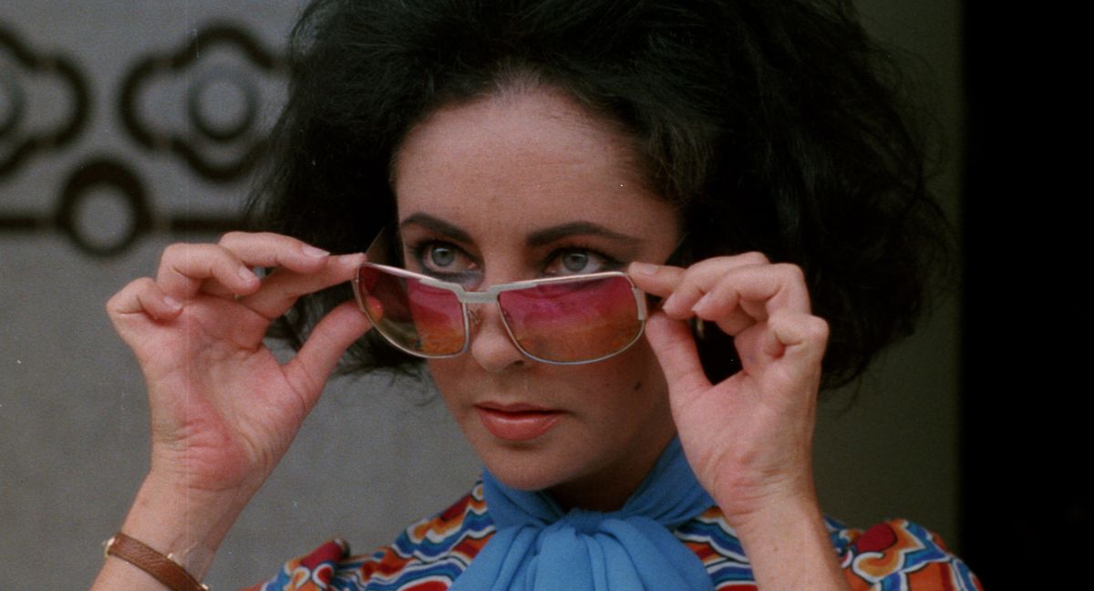 Elizabeth Taylor si abbassa gli occhiali da sole colorati fino al ponte del naso con entrambe le mani in Identikit.