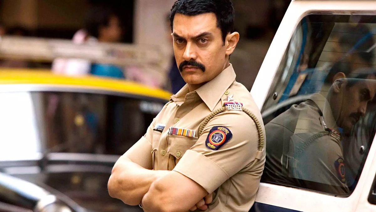 Aamir Khan, che indossa un'uniforme della polizia e sfoggia folti baffi, incrocia le braccia mentre si appoggia alla portiera di un'auto a Talaash.