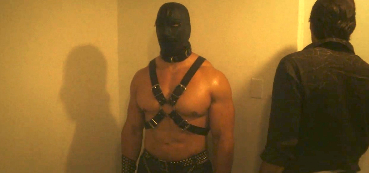 un uomo muscoloso con una maschera bdsm in pelle e un'imbracatura in piedi accanto a una porta, mentre un altro uomo apre la porta, apparentemente ignaro del ragazzo muscoloso