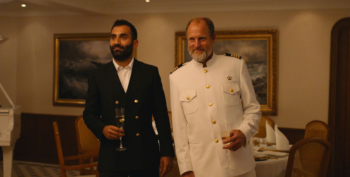 Il capitano dello yacht senza nome (Woody Harrelson) in Triangle of Sadness, in uniforme completamente bianca, è in piedi accanto a un ospite sorridente e vestito di nero sullo yacht durante la cena del capitano