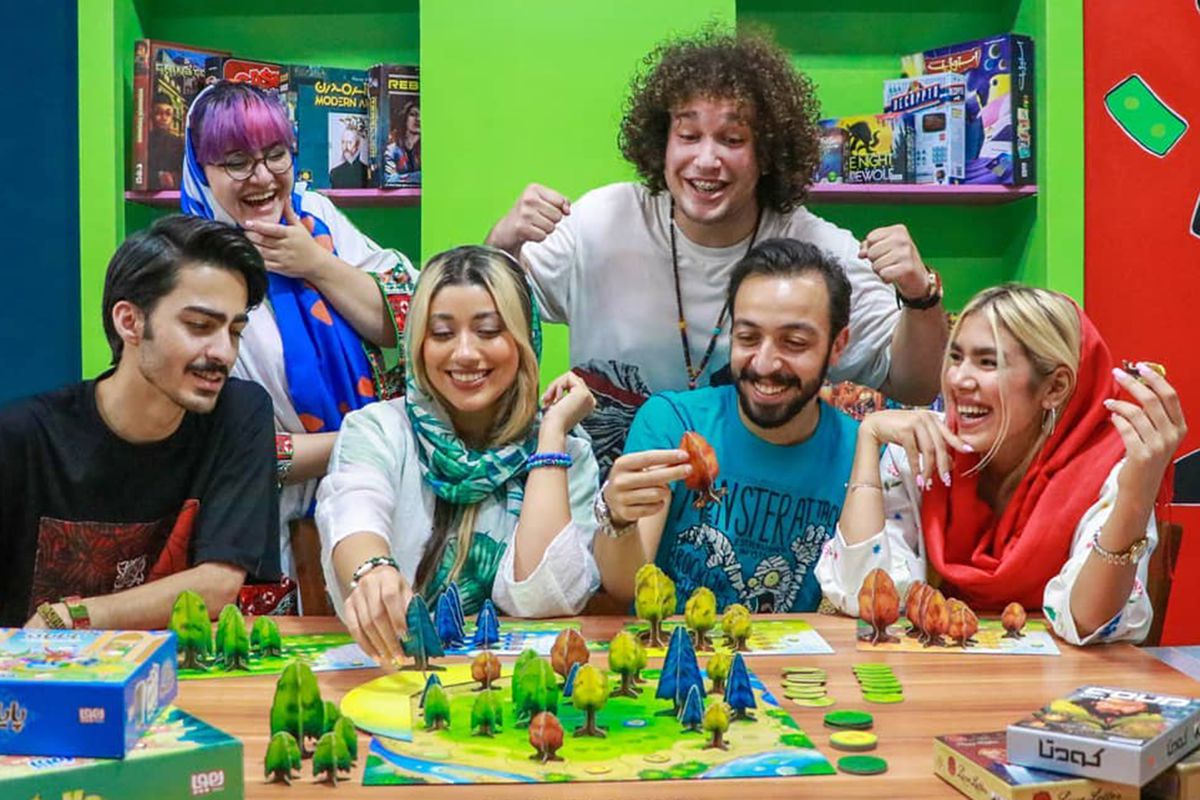 Sei giovani iraniani con una copia di Photosintesi di Blue Orange. Il tavolo è pieno di piccoli alberi e tutti ridono.