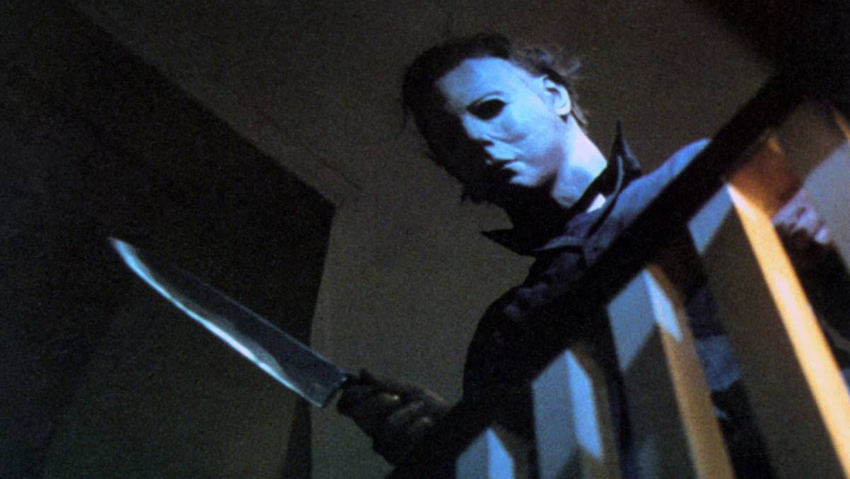 Michael Myers nella sua maschera bianca originale si sporge su una ringhiera delle scale e tiene un coltello in modo minaccioso ad Halloween.
