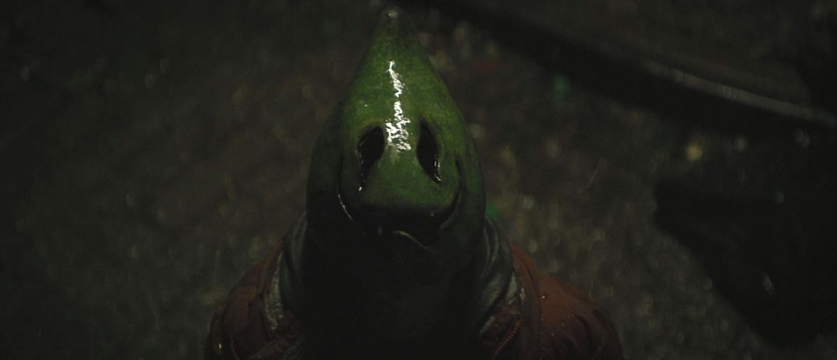 Un rapinatore con una maschera a forma di avocado verde lucido con bocca e occhi irregolari da spaventapasseri si ferma sotto la pioggia e guarda il Bat-Segnale in The Batman