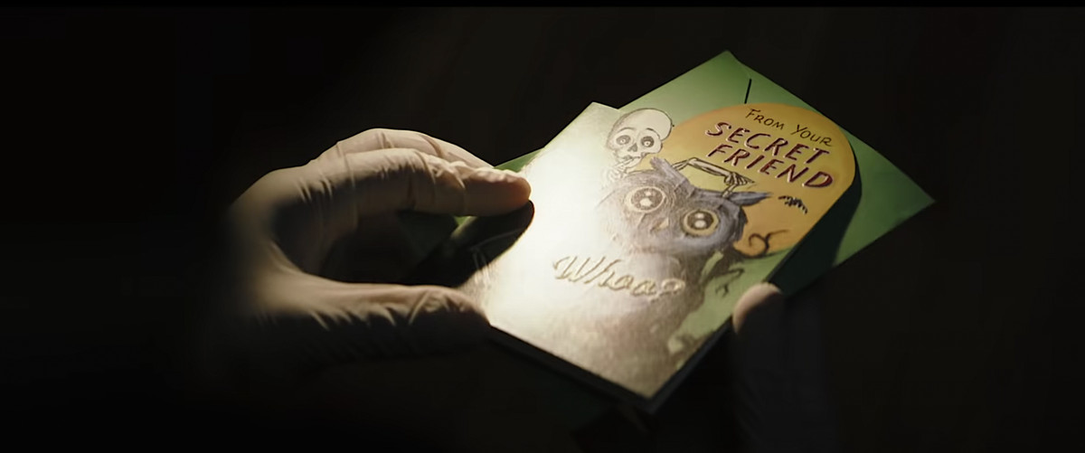 Una mano che indossa un guanto della polizia in lattice tiene un biglietto di auguri di Halloween con uno scheletro in piedi dietro un gufo e indicandolo mentre tiene in mano un