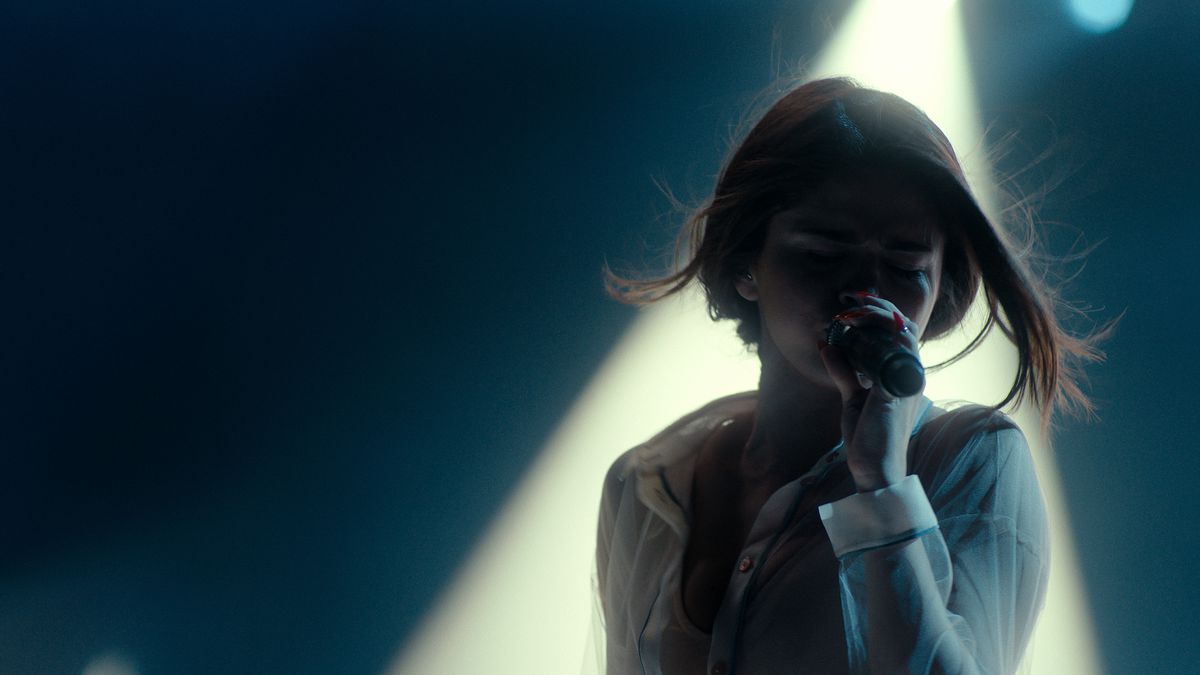 Una donna (Selena Gomez) tiene un microfono in una mano e canta mentre è illuminata da un raggio di luce sullo sfondo.