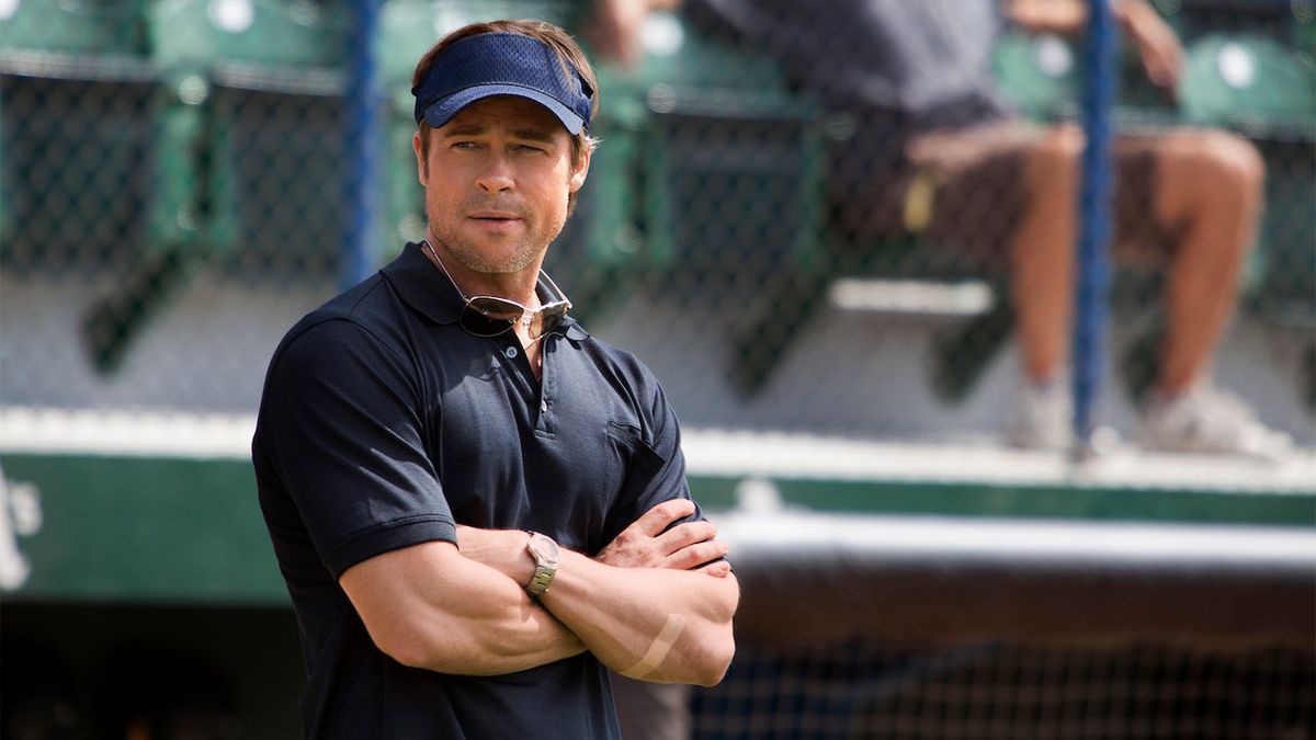 Un uomo con una polo blu navy (Brad Pitt) che indossa un cappello blu mentre è in piedi sul lato di un campo da baseball.