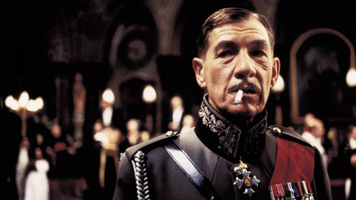Un uomo in abiti militari (Ian McKellen) con una sigaretta in bocca in una sala sontuosa con candele e folla sullo sfondo.