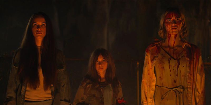 Tre donne stanno una accanto all'altra, con la donna all'estrema destra inzuppata di sangue.