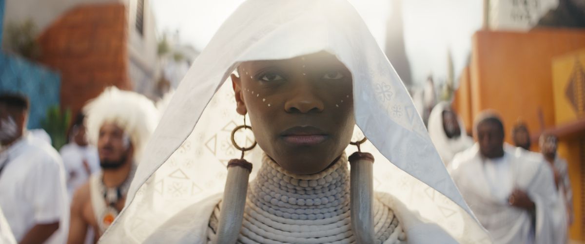 Una giovane donna (Letitia Wright) adornata con uno scialle funebre bianco con lunghi orecchini d'avorio.