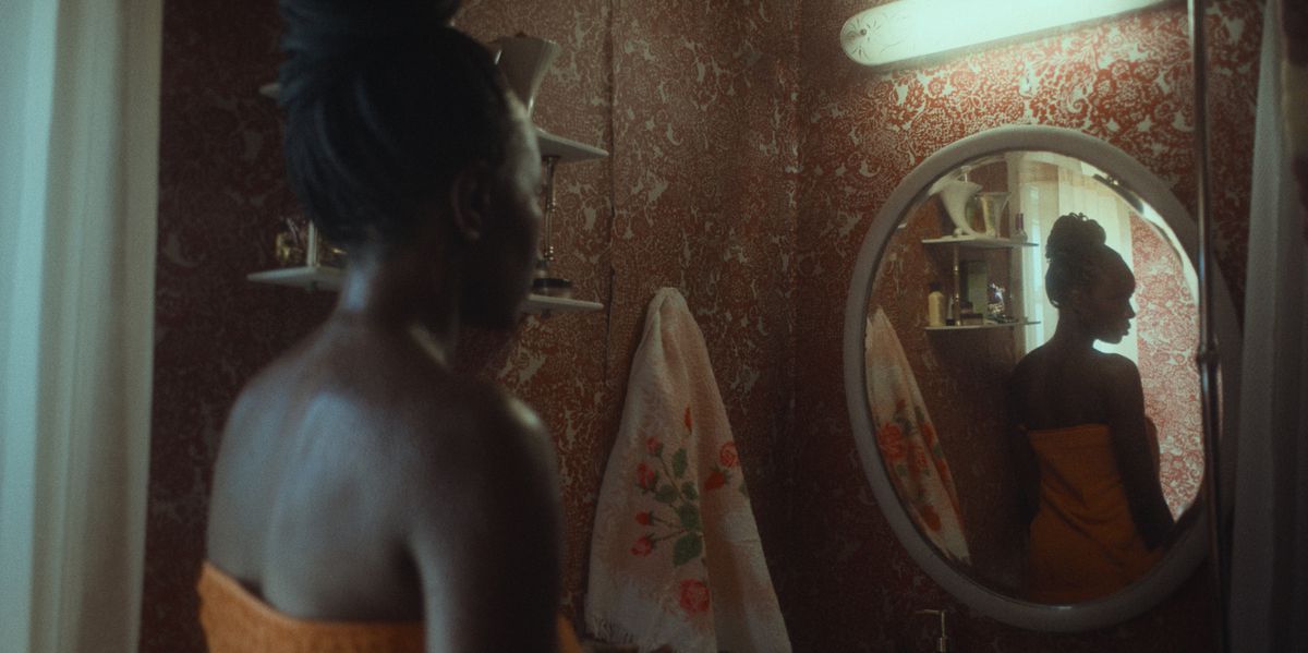 Aisha (Anna Diop), una donna dalla pelle scura che indossa un asciugamano arancione brillante, si esamina allo specchio in una stanza buia in Nanny