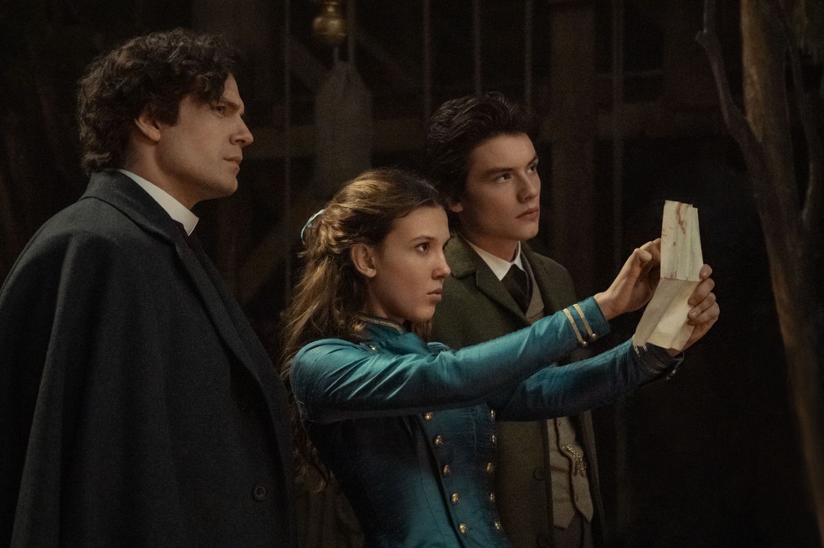 Henry Cavill nei panni di Sherlock Holmes, Millie Bobby Brown nei panni di Enola Holmes, Louis Partridge nei panni di Tewkesbury in Enola Holmes 2. Enola alza un pezzo di carta e lo indica mentre gli altri guardano.