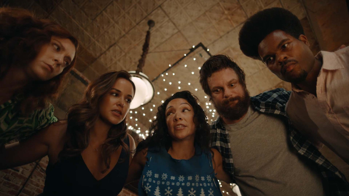 Una lunga inquadratura di un cerchio di uomini e donne rannicchiati insieme e che parlano intensamente, il soffitto di un bar visibile sopra.