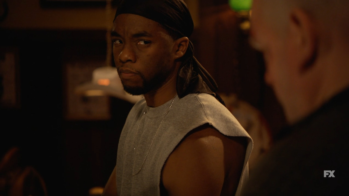 Chadwick Boseman indossa una maglietta senza maniche e un durag in Justified, fissando un uomo accanto a lui.