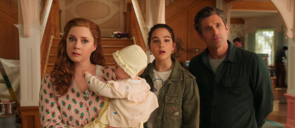 Una donna dai capelli rossi con una camicia a fiori rosa (Amy Adams) tiene in braccio un bambino accanto a una ragazza con un cappotto verde (Morgan Philip) e un uomo (Patrick Dempsey) mentre si trova di fronte a una scala e a un soggiorno.