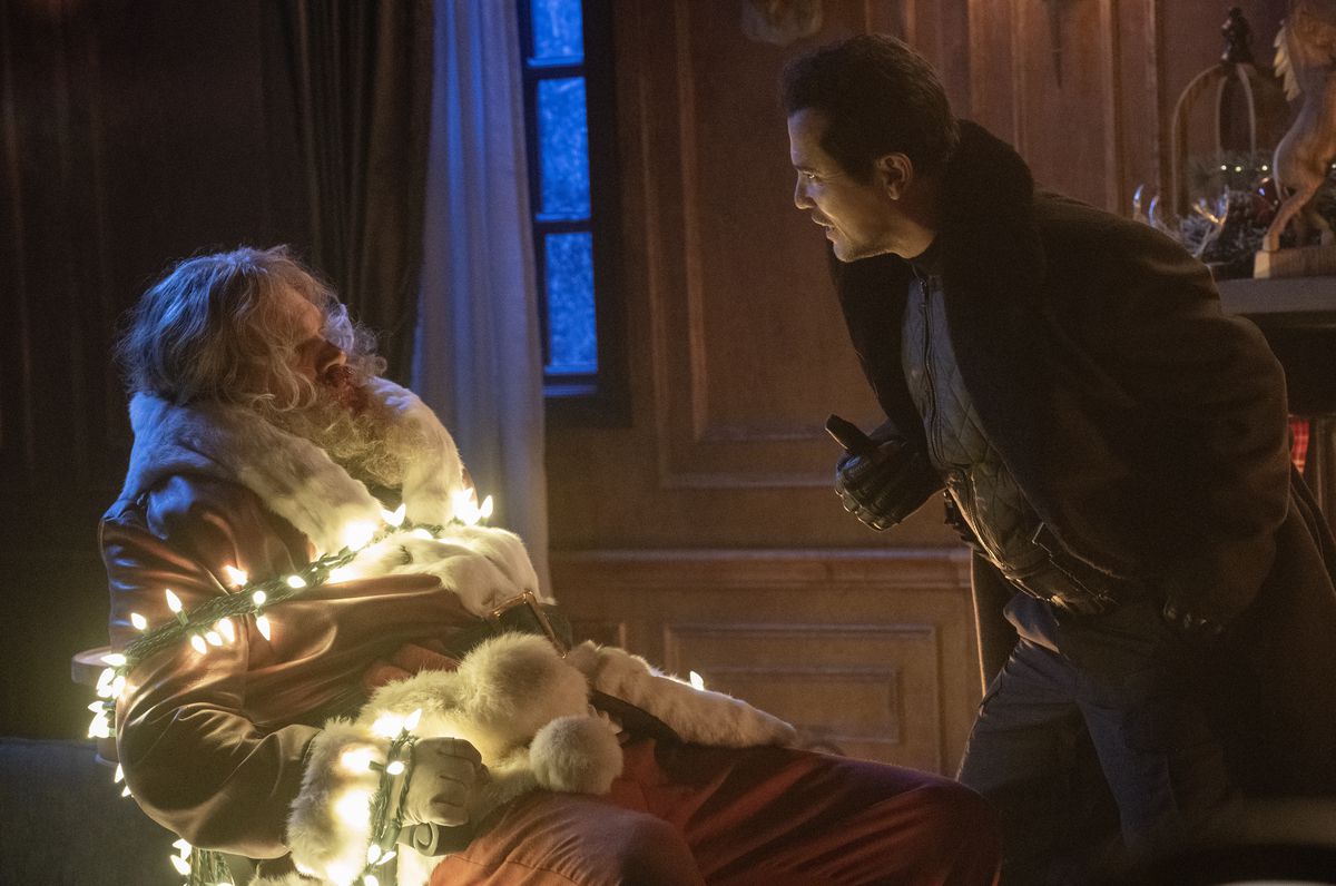 Scrooge (John Leguizamo) si china minacciosamente su Babbo Natale (David Harbour), che è legato a una sedia con una serie di luci natalizie bianche accese, in Violent Night