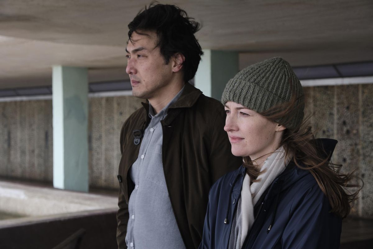 Un uomo che indossa un cappotto marrone (Takehiro Hira) è in piedi accanto a una donna con un berretto di maglia grigio (Kelly Macdonald) e una giacca blu, i capelli spazzati di lato dal vento.