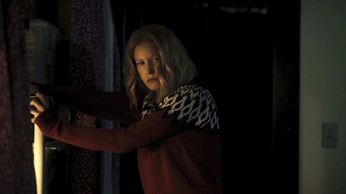 Una donna con un maglione rosso festivo (Anna Gunn) si appoggia a un caminetto e si guarda alle spalle con un'espressione di disgusto.