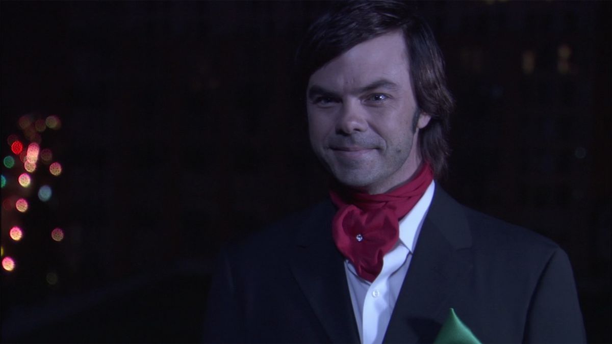 Un ragazzo stupido con i capelli mossi e un ascot rosso in piedi accanto a un albero di Natale illuminato in Chasing Christmas (2005)