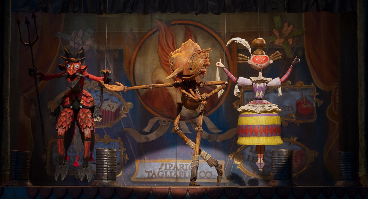 Un burattino di legno si inchina sul palco accanto a due pupazzi dipinti come un demone e una donna in costume.