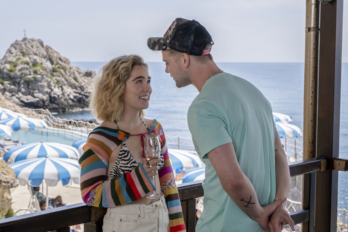 Haley Lu Richardson nel ruolo di Portia in White Lotus. Sta parlando con un giovane fuori in un bar con patio affacciato sull'oceano. Indossa un top bolero in maglia arcobaleno con un top bikini zebrato.