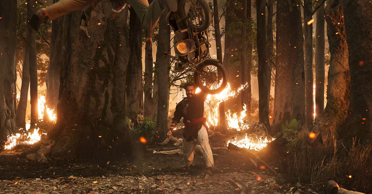 Un uomo indiano corpulento lancia una motocicletta attraverso una foresta infuocata con una mano in RRR
