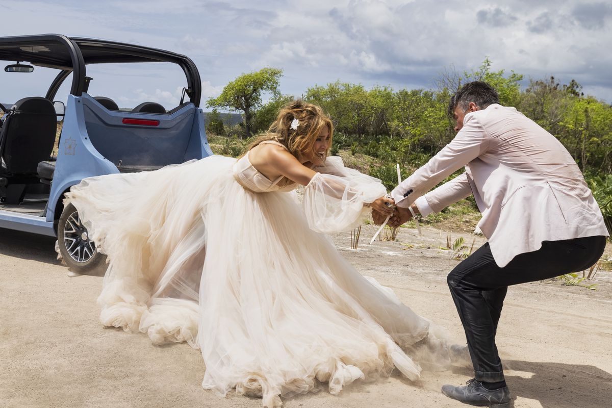 Josh Duhamel si aggrappa alle braccia di Jennifer Lopez mentre si allunga dal retro di un'auto in Shotgun Wedding. Entrambi indossano abiti da sposa.
