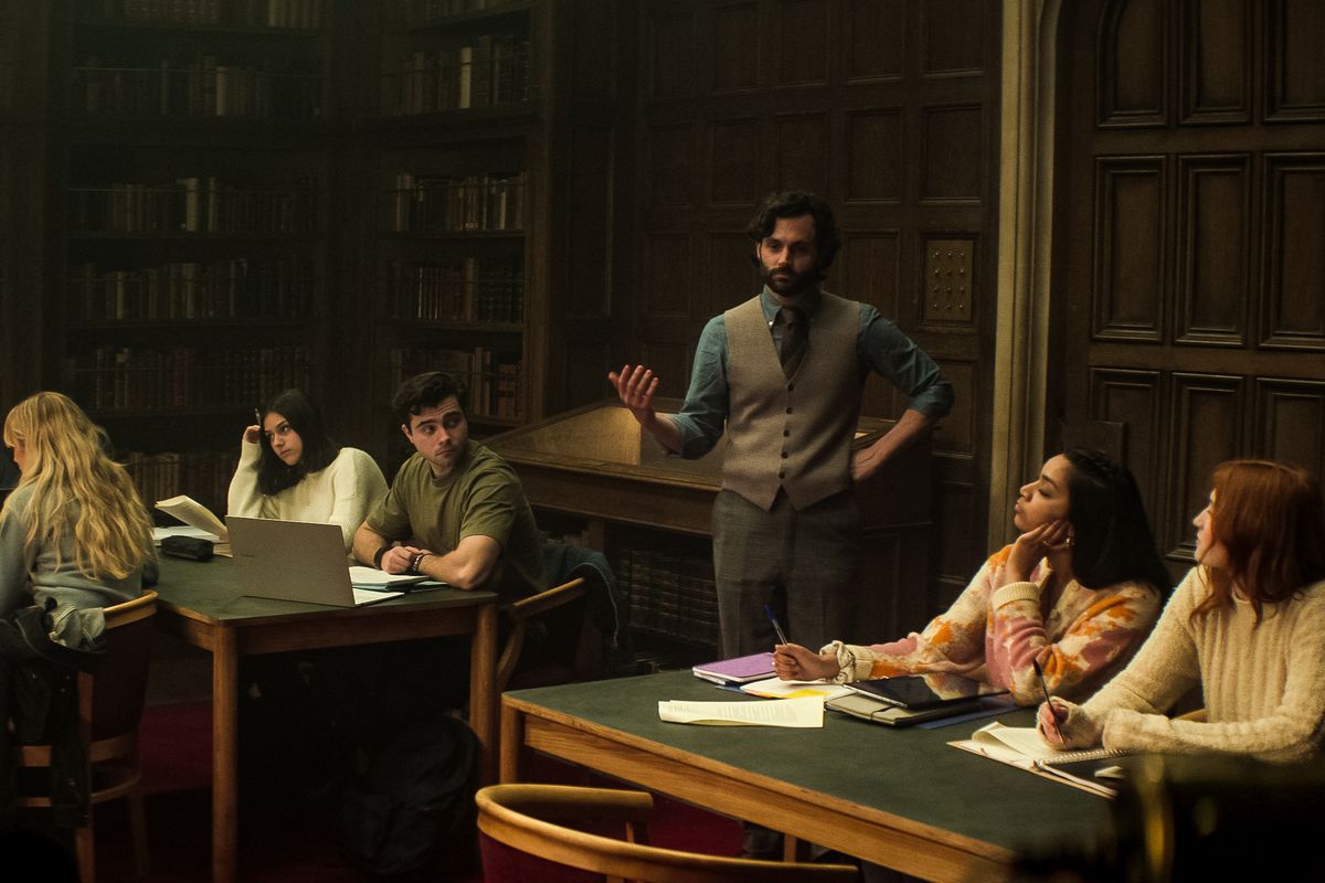 Penn Badgley indossa un giubbotto e parla con un gruppo di studenti in una biblioteca nella quarta stagione di You.