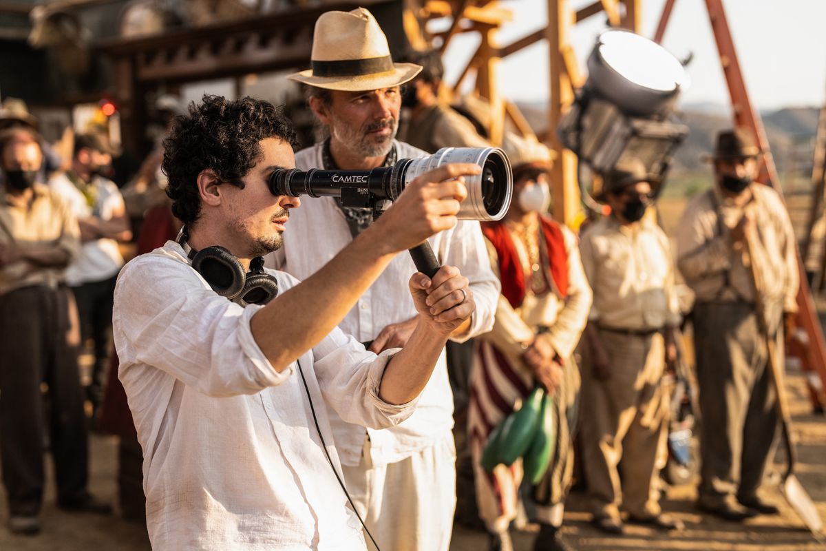 Damien Chazelle tiene in mano il mirino di un regista sul set di Babylon mentre il direttore della fotografia Linus Sandgren è in piedi dietro di lui con un cappello con un gruppo di comparse vestite d'epoca in piedi sullo sfondo vicino a un finto set cinematografico
