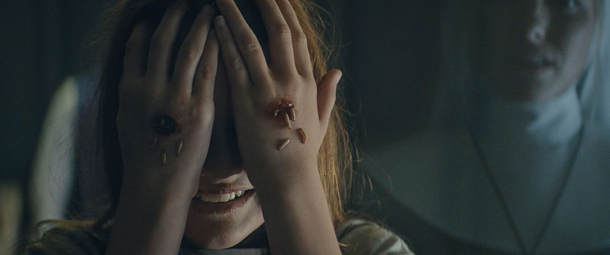 Una giovane ragazza sorride mentre si copre gli occhi con i palmi delle mani, mentre i vermi strisciano fuori dalle ferite visibili delle stigmate sul dorso delle sue mani e una giovane suora sembra inorridita sullo sfondo.