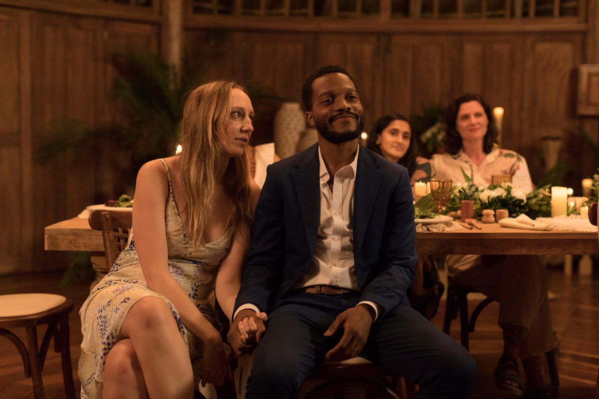 Una donna in abito bianco (Jillian Bell) siede accanto a un principale sorridente che indossa un blazer blu e pantaloni grigi (Utkarsh Ambudkar) seduto a un tavolo da pranzo.