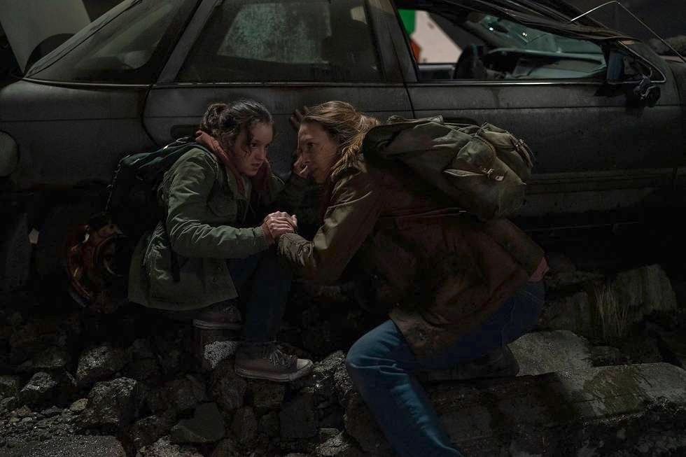 Ellie (Bella Ramsey) e Tess (Anna Torv) accovacciate dietro un'auto decrepita in un fotogramma di The Last of Us