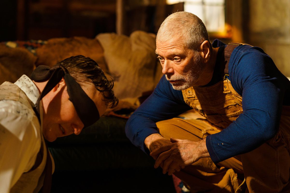 Un uomo anziano (Stephen Lang) con una camicia blu a maniche lunghe e una tuta gialla si accovaccia davanti a un uomo bendato (Marc Senter).