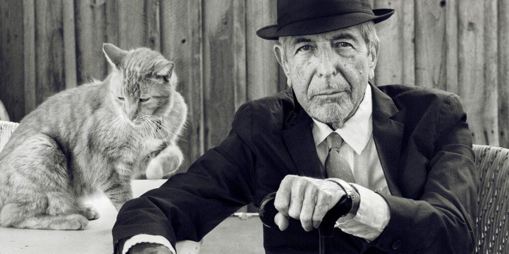 Una foto d'archivio in bianco e nero di Leonard Cohen in abito scuro seduto su una sedia con in mano un bastone accanto a un tavolo con sopra un gatto.