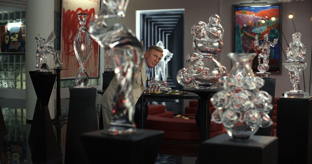 Il detective Benoit Blanc (Daniel Craig) scruta attraverso una serie di sculture di vetro su piedistalli dall'aspetto ricky in una stanza piena di opere d'arte in Glass Onion