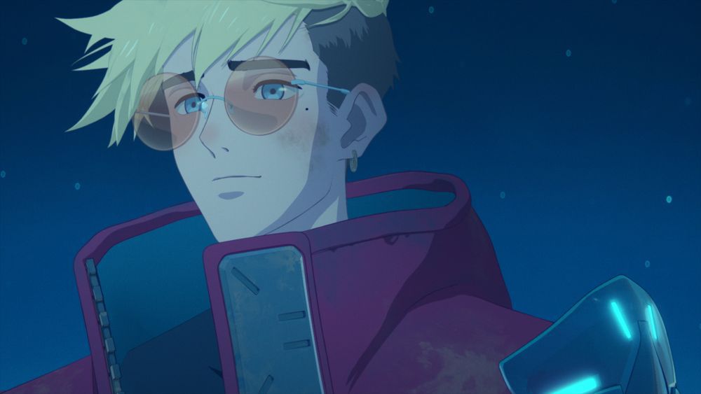 Un uomo anime dai capelli gialli (Vash the Stampede) con un cappotto rosso e occhiali circolari arancioni che sorride e fissa in lontananza.