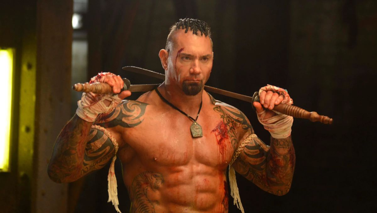 Dave Bautista, a torso nudo e con del sangue addosso, tiene due spade sulle spalle e ha delle bende intorno alle mani in Kickboxer: Vengeance