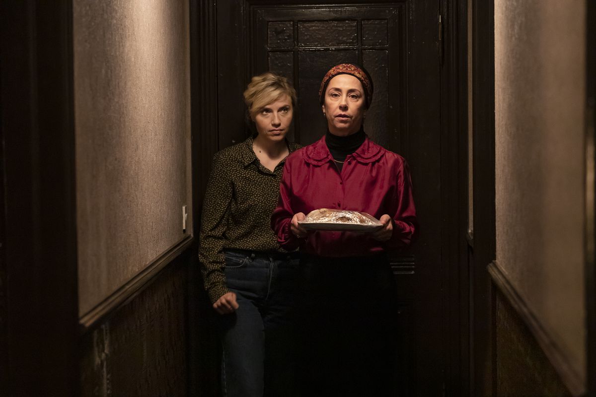 Due persone escono da una porta. Uno indossa un foulard e una camicia rossa e porta un piatto di cibo. L'altro guarda l'altra persona da dietro, con capelli biondi e jeans, in Attachment.