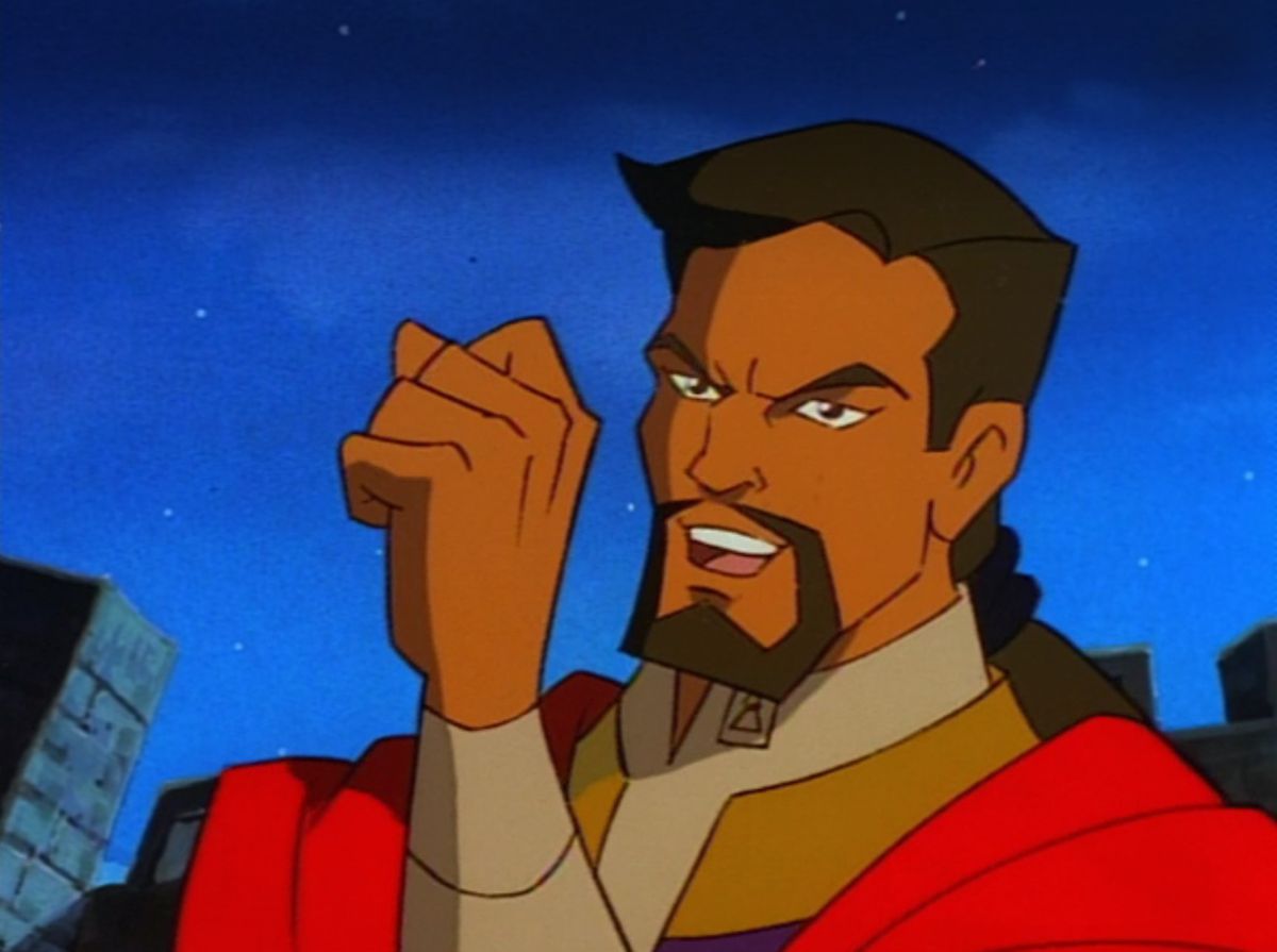 Un primo piano di un uomo con pizzetto marrone e coda di cavallo in una veste rossa e oro che tiene il pugno davanti alla mano e parla.