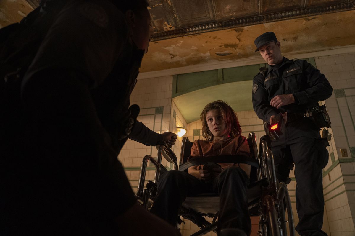 Un ufficiale della FEDRA in piedi sopra un bambino legato a una sedia a rotelle