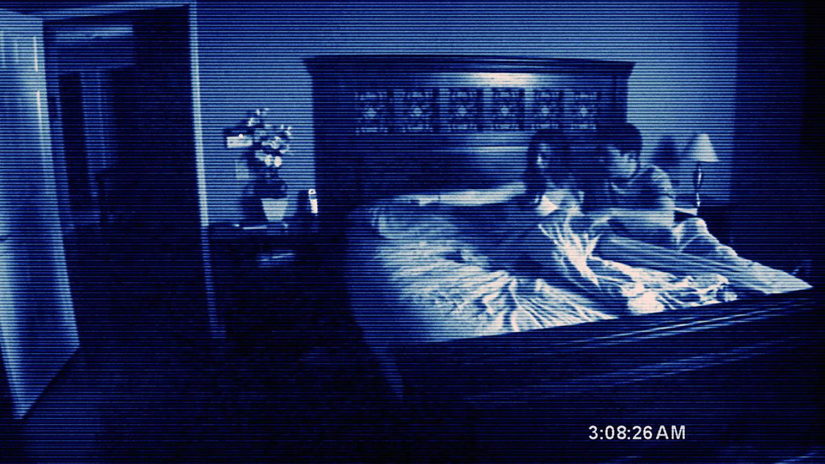 Una coppia si rannicchia nel letto, indicando con timore qualcosa fuori dallo schermo, in un'immagine sgranata e a bassa risoluzione della telecamera da Paranormal Activity del 2007