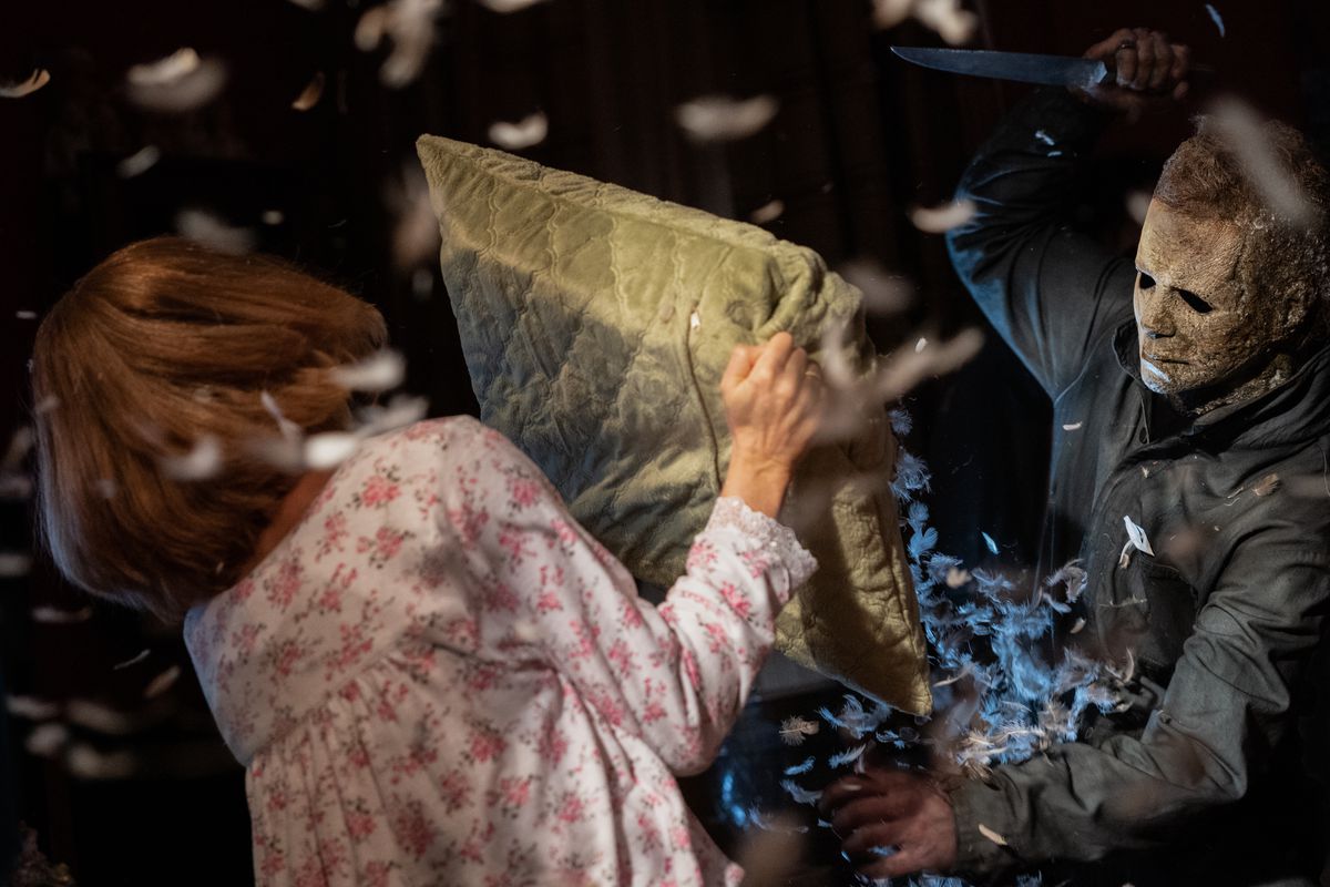 Una persona dai capelli rossi in pigiama con le spalle rivolte alla telecamera cerca di respingere un Michael Myers armato di coltello con un cuscino - davvero, un cuscino - e piume volano ovunque in una ripresa di Halloween Ends