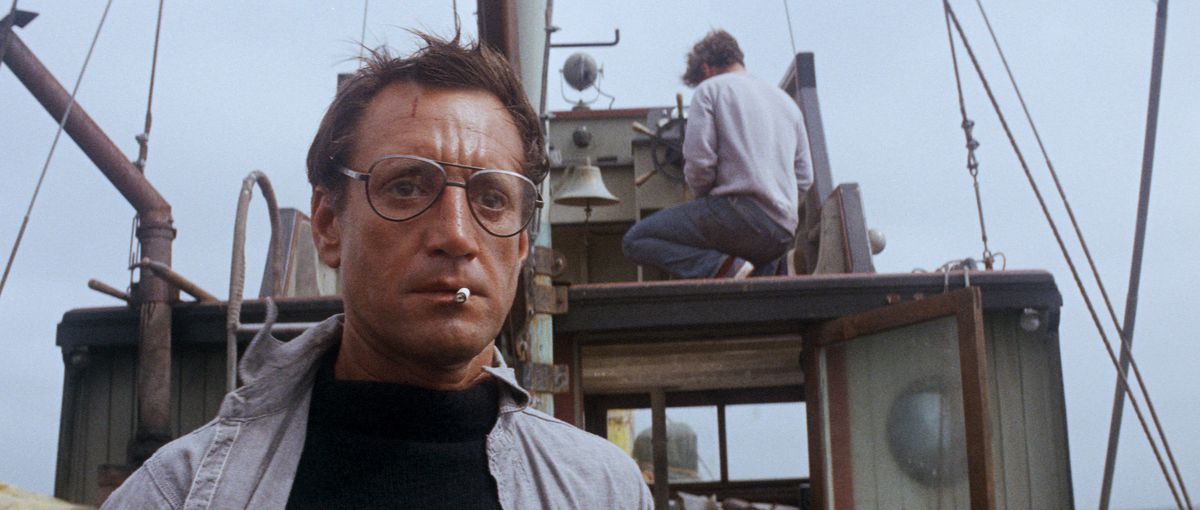 Roy Scheider nei panni di Hooper che indossa grandi occhiali e fuma una sigaretta, fissa scioccato il retro di una barca in Jaws