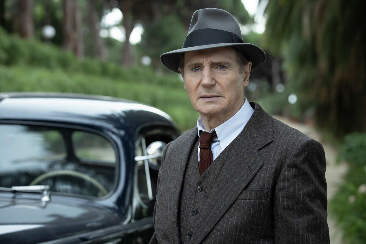 Un uomo (Liam Neeson) in piedi in una zona boscosa di fronte a un abito da berlina scuro con un abito gessato marrone, cravatta rosso scuro e un fedora grigio.