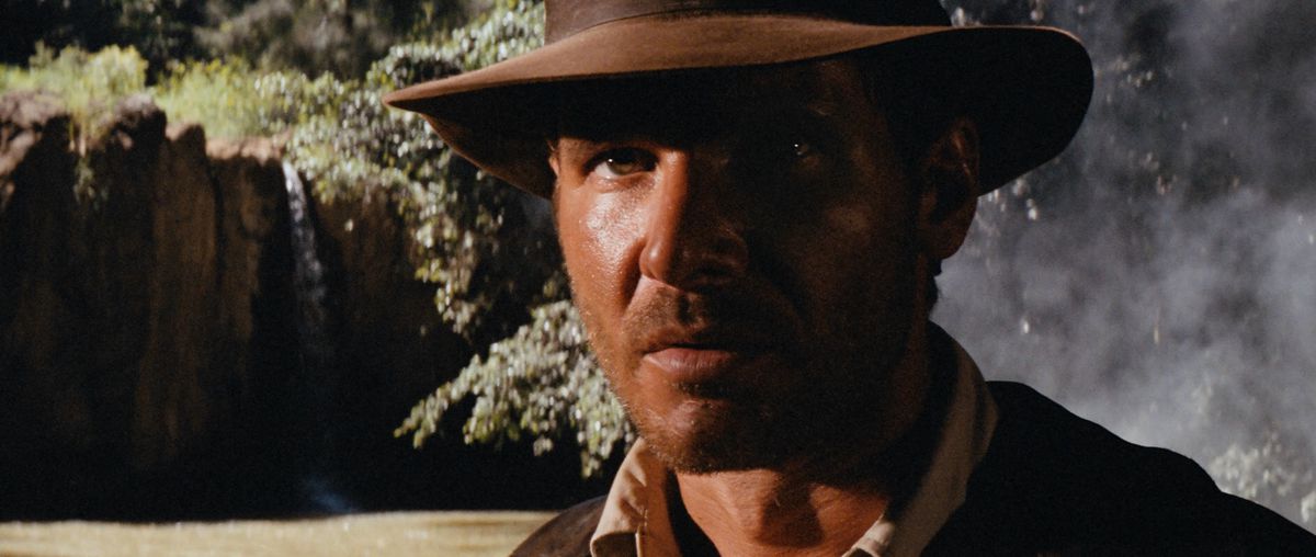 Indiana Jones si trova vicino a un fiume fangoso e alla giungla e guarda fuori dallo schermo mentre è avvolto nell'ombra in I predatori dell'arca perduta