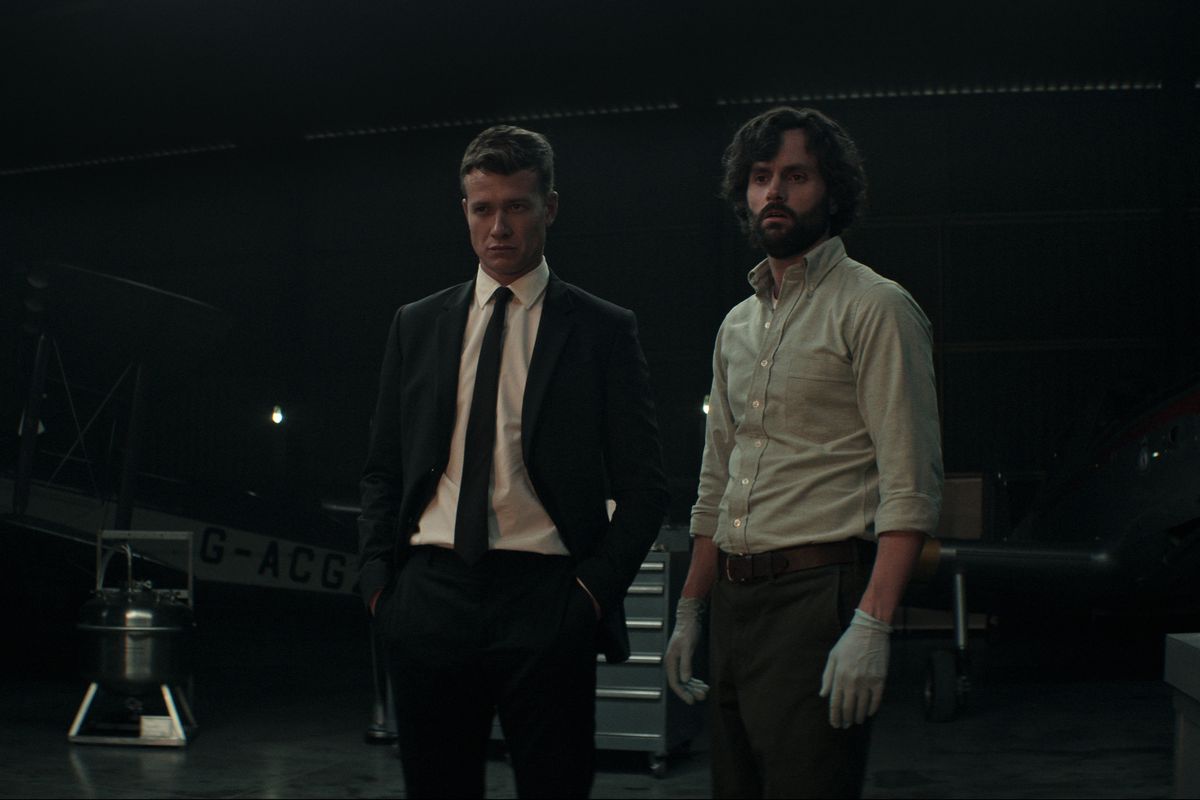 Rhys in abito nero e Joe in camicia con maniche rimboccate e guanti di lattice, stanno uno accanto all'altro in uno scantinato buio nella stagione 4 di You