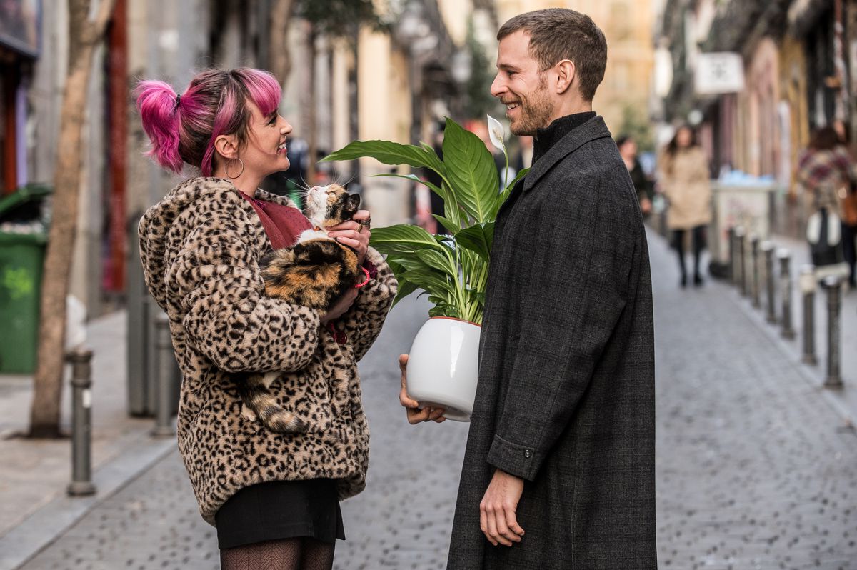 Una coppia sta insieme per strada in Amore al primo bacio. A sinistra, una persona con i capelli rosa indossa un cappotto leopardato e tiene in braccio un gatto. A destra, una persona indossa un lungo cappotto nero e tiene in mano una pianta in vaso