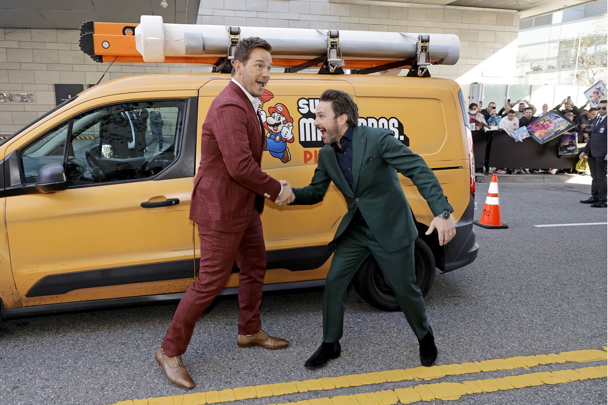Chris Pratt e Charlie Day si trovano davanti a un minivan a tema, alla premiere di The Super Mario Bros LA. Pratt indossa un abito rosso intenso e Day indossa un abito verde intenso, ei due si stringono la mano con espressioni buffe sui loro volti.