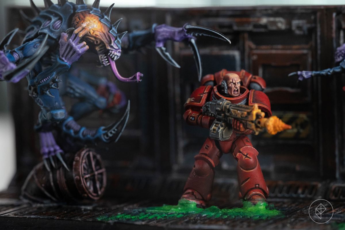 Two tyrranids flank a red-armored Space Marine miniature. The floor of this space hulk is covered with a bright green ichor.